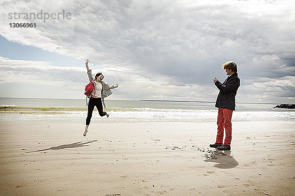 Mann fotografiert springende Frau  während er am Strand auf Sand steht