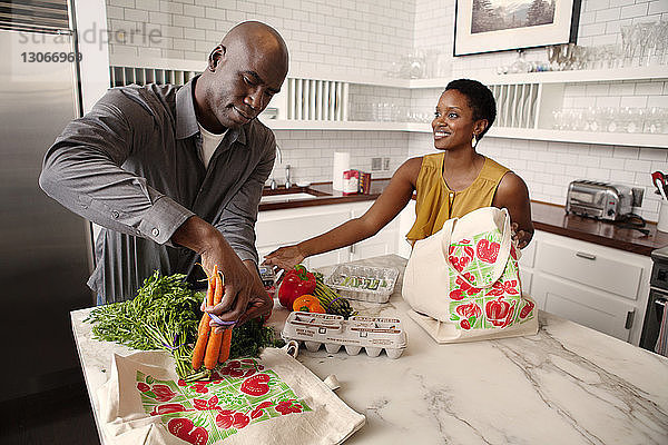 Frau sieht Ehemann an  wie er Gemüse auf Kücheninsel legt