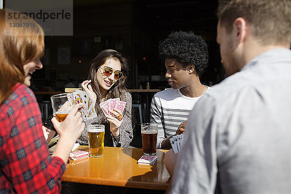 Lächelnde Freunde spielen Karten  während sie in der Brauerei Bier am Tisch trinken