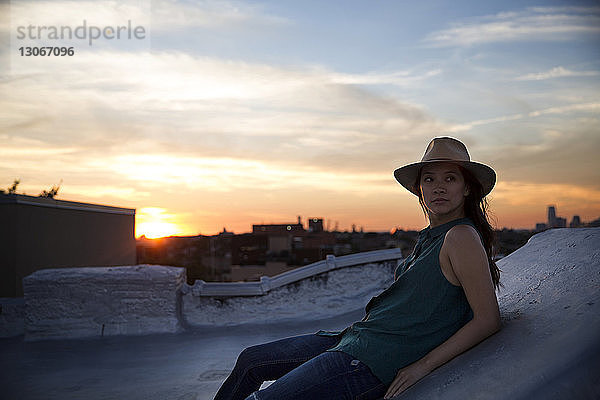 Nachdenkliche Frau lehnt bei Sonnenuntergang auf Stützmauer gegen den Himmel