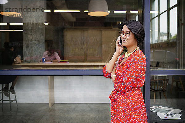 Frau spricht mit Smartphone  während sie sich am Büroeingang anlehnt