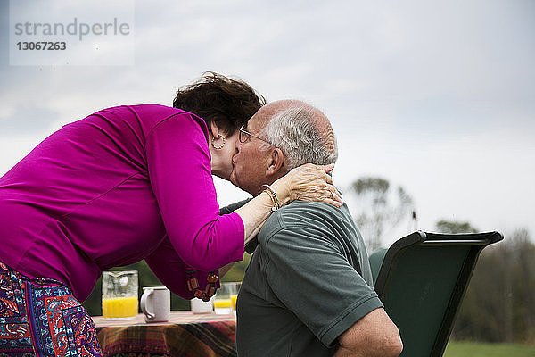 Seitenansicht eines älteren Mannes  der seine Frau küsst  während er am Tisch gegen den Himmel sitzt