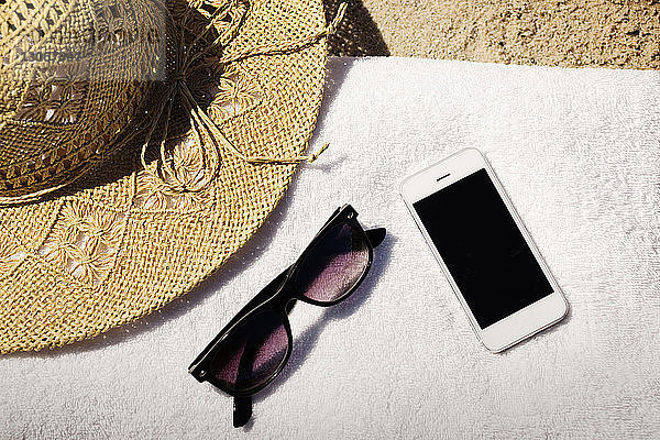 Draufsicht auf Sonnenbrille und Mobiltelefon mit Strohhut auf einem Handtuch am Strand
