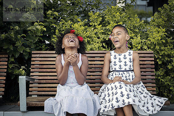 Fröhliche Schwestern sitzen auf Holzbank gegen Pflanzen in der Stadt
