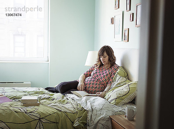 Schwangere Frau liest Buch im Sitzen im Bett