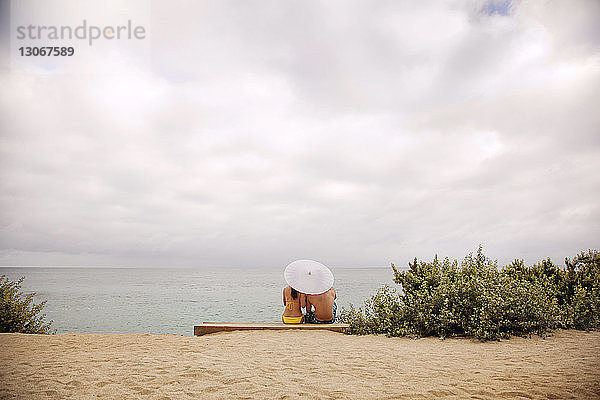 Rückansicht eines am Strand sitzenden Paares mit Sonnenschirm vor bewölktem Himmel