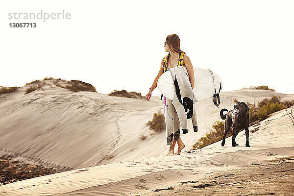 Frau trägt Surfbrett beim Spaziergang mit Hund auf Sand vor klarem Himmel