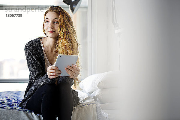 Frau mit Tablet-Computer schaut weg  während sie zu Hause auf dem Bett sitzt