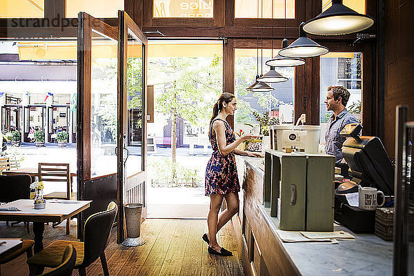 Weiblicher Kunde hält Kaffeetasse in der Hand  während er sich mit dem Besitzer am Tresen des Cafés unterhält