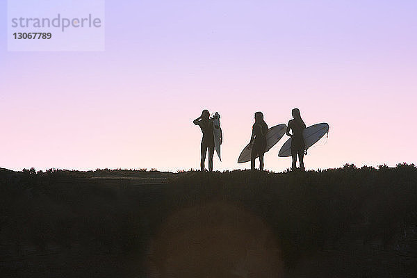 Silhouette von Freundinnen  die ein Surfbrett halten  während sie bei Sonnenuntergang auf dem Feld stehen
