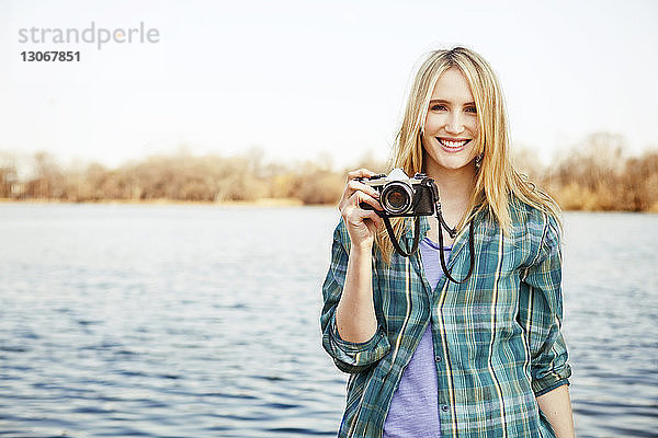Porträt einer Frau  die am Seeufer stehend die Kamera hält