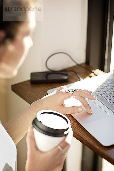 Frau hält Kaffeetasse  während sie im Café einen Laptop benutzt
