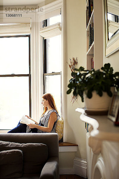 Frau liest Buch  während sie zu Hause auf einem Fensterplatz in einer Nische sitzt