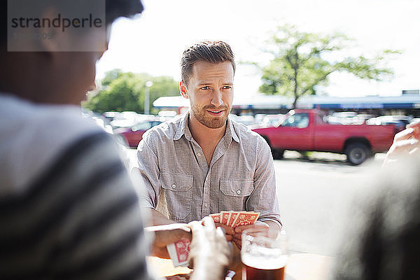 Mann schaut Freund an  während er in einer Brauerei am Tisch Karten gegen Fenster spielt