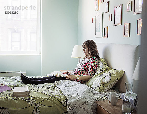 Schwangere Frau liest zu Hause auf dem Bett sitzend ein Buch