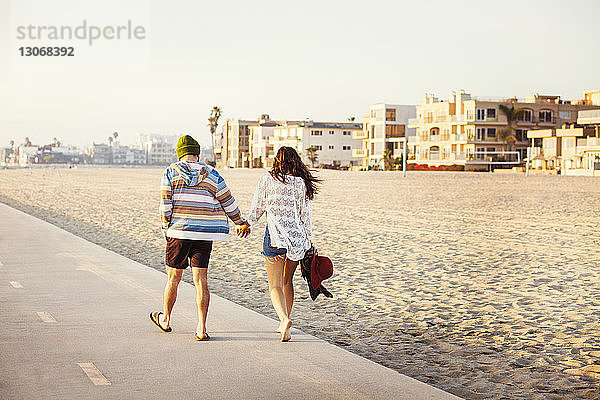 Rückansicht eines Paares  das sich an sonnigen Tagen an den Händen hält und am Strand spazieren geht
