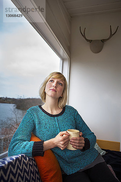 Nachdenkliche Frau trinkt Kaffee  während sie zu Hause durchs Fenster schaut