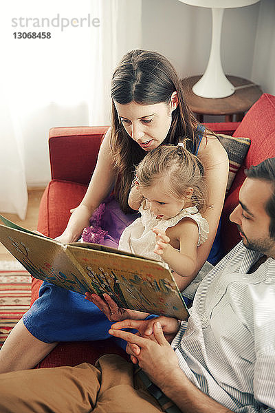 Mann und Frau zeigen einem Mädchen ein Bilderbuch  während sie zu Hause auf dem Sofa sitzen