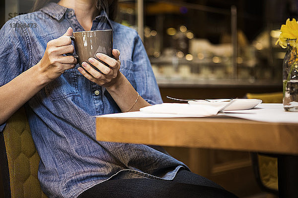 Mittelteil einer Frau mit Kaffeetasse in der Hand  die in einem Café am Tisch sitzt