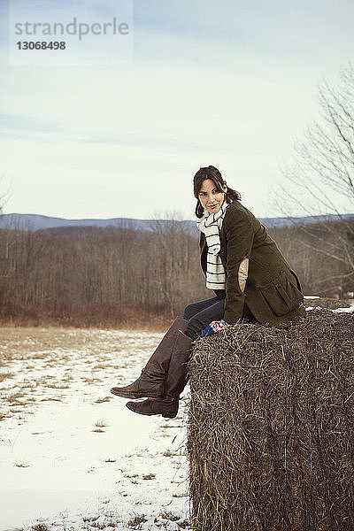 Frau schaut weg  während sie im Winter auf Heuballen gegen den Himmel sitzt