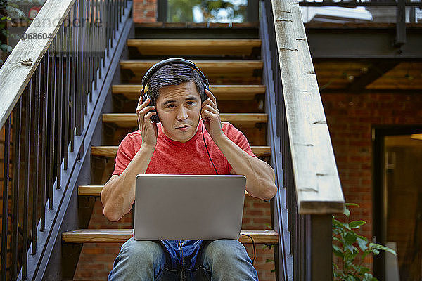Mann hört Musik über Kopfhörer von einem Laptop-Computer  während er auf einer Treppe im Hof sitzt