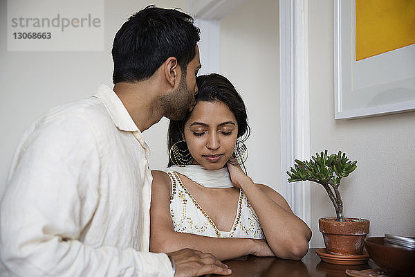 Mann küsst Frau auf die Stirn  während er zu Hause am Tisch steht