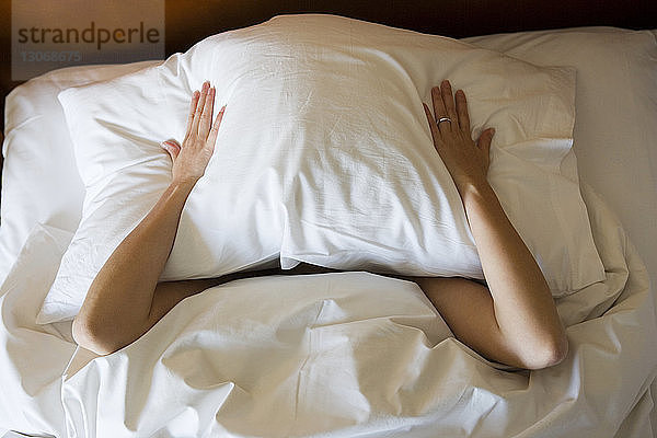 Hochwinkelansicht einer Frau  die das Gesicht mit einem Kissen bedeckt  während sie auf dem Bett liegt