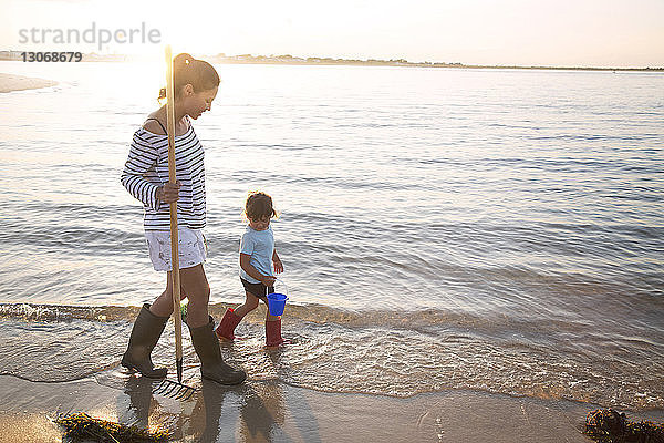 Mutter und Tochter gehen bei Sonnenschein mit Harke und Eimer am Meeresufer spazieren