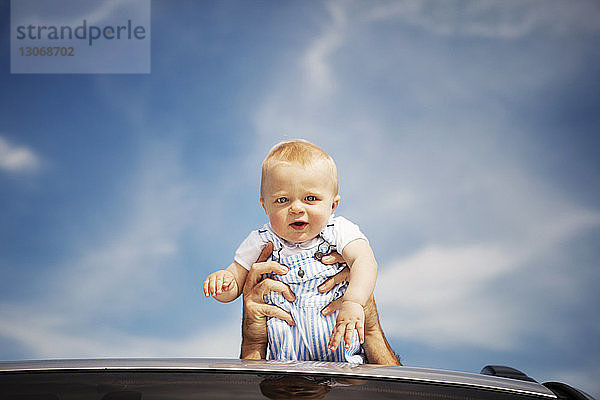 Porträt eines neugierigen kleinen Jungen  der vom Vater über das Autodach gegen den Himmel getragen wird