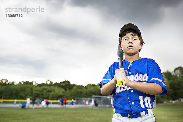 Porträt eines Jungen mit Baseballschläger auf einem Feld vor bewölktem Himmel stehend