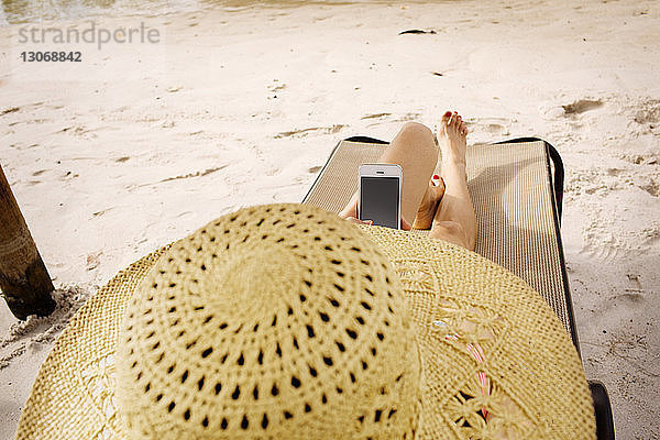 Hochwinkelansicht einer Frau  die ein Smartphone benutzt  während sie sich auf einem Liegestuhl am Strand entspannt