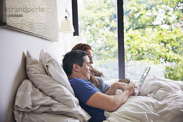 Seitenansicht eines Paares  das einen Tablet-Computer benutzt  während es zu Hause auf dem Bett liegt