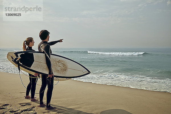Paar schaut aufs Meer  während es ein Surfbrett am Strand trägt