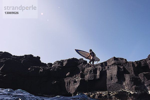 Frau trägt Surfbrett  während sie auf Felsen gegen den klaren Himmel läuft