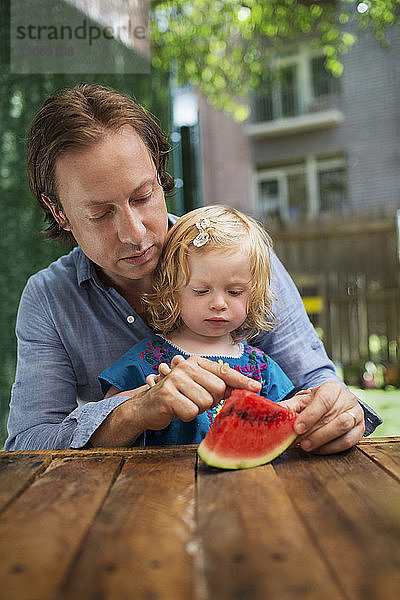 Vater zeigt der Tochter eine Wassermelone  während er im Garten am Tisch sitzt