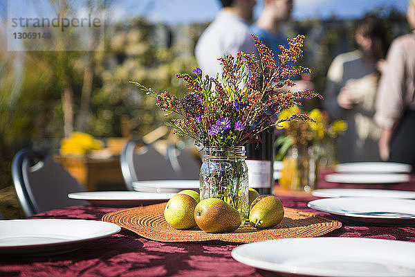 Blumenvase und Früchte auf einem Tisch im Freien mit Freunden im Hintergrund bei einer Gartenparty