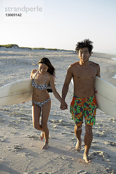 Paar mit Surfbrettern  die sich beim Strandspaziergang am Ufer an den Händen halten