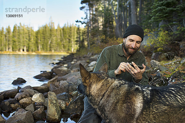 Mann mit deutschem Schäferhund benutzt Mobiltelefon am Seeufer