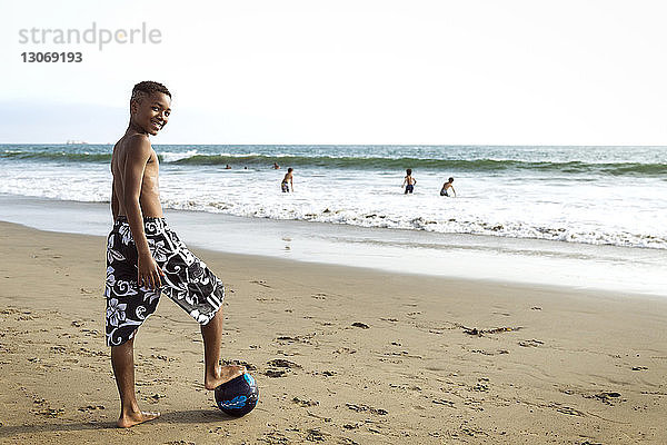 Porträt eines Jungen  der mit einem Bein auf einem Fussball am Strand steht