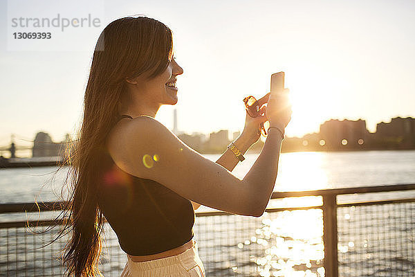 Glückliche Frau fotografiert mit Handy am Fluss bei Sonnenuntergang
