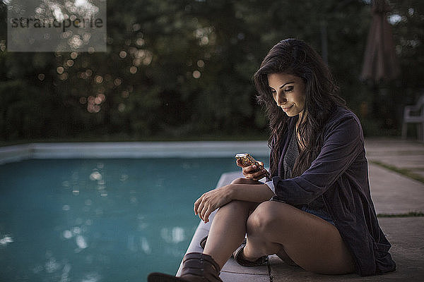 Frau benutzt Mobiltelefon  während sie bei Sonnenuntergang am Pool sitzt