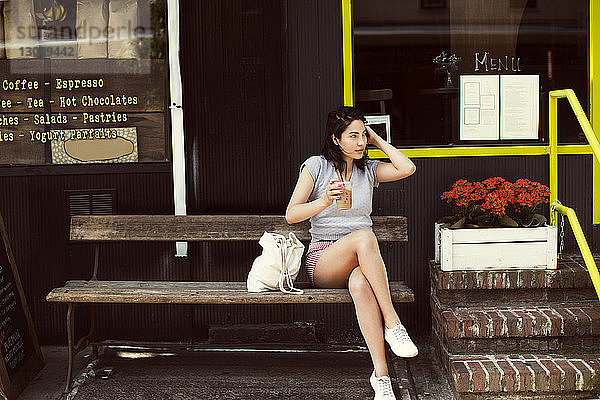 Nachdenkliche Frau trinkt Kaffee  während sie auf einer Bank im Bürgersteig-Café in der Stadt sitzt