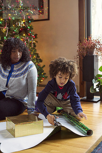 Mutter sieht Sohn an  der zu Weihnachten zu Hause Geschenkpapier schneidet