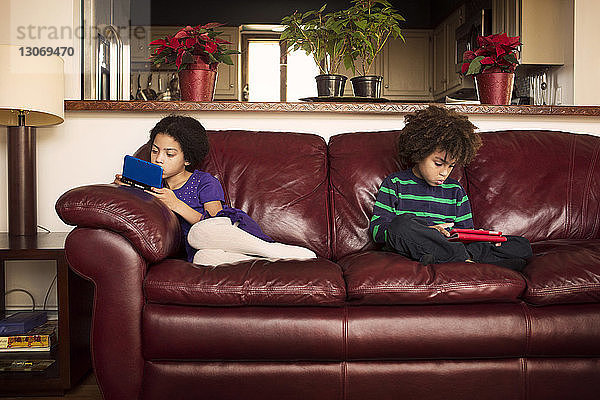 Bruder und Schwester nutzen Technologien  während sie zu Hause auf einem Ledersofa sitzen