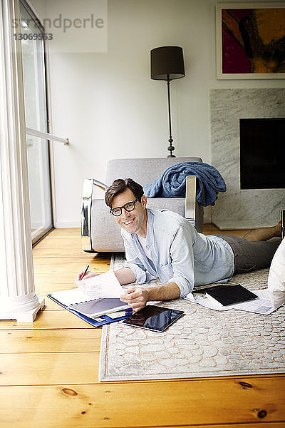 Lächelnder Mann erledigt Papierkram per Tablet-Computer  während er zu Hause auf dem Boden liegt