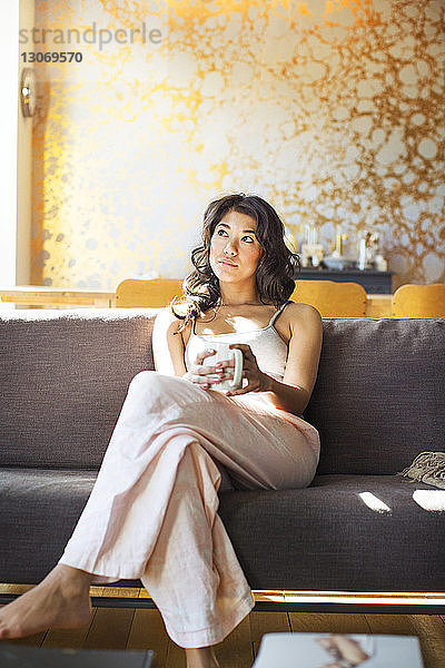 Nachdenkliche Frau hält Kaffeetasse und schaut weg  während sie sich auf dem Sofa entspannt
