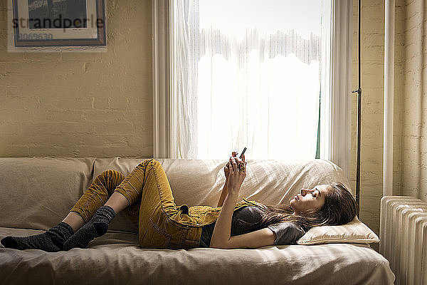 Seitenansicht einer Frau  die ein Smartphone benutzt  während sie sich zu Hause auf dem Sofa entspannt