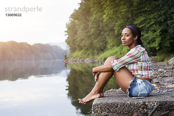 Seitenansicht einer Frau  die weg schaut  während sie auf einem Felsen gegen einen See im Wald sitzt