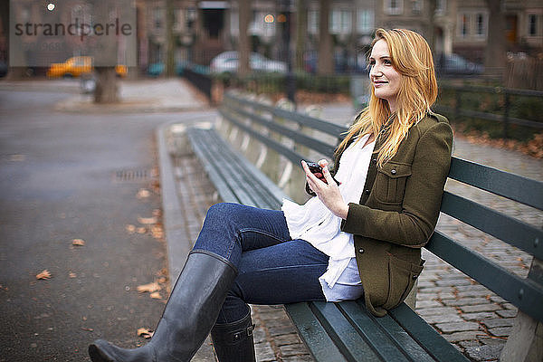 Frau mit Mobiltelefon schaut weg  während sie auf einer Bank sitzt