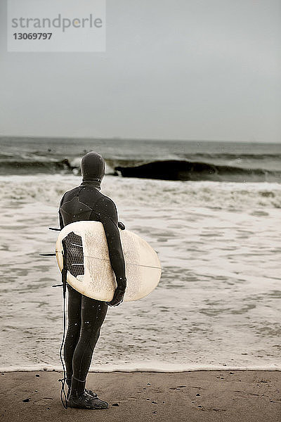 Rückansicht eines Mannes  der ein Surfbrett trägt  während er am Strand steht